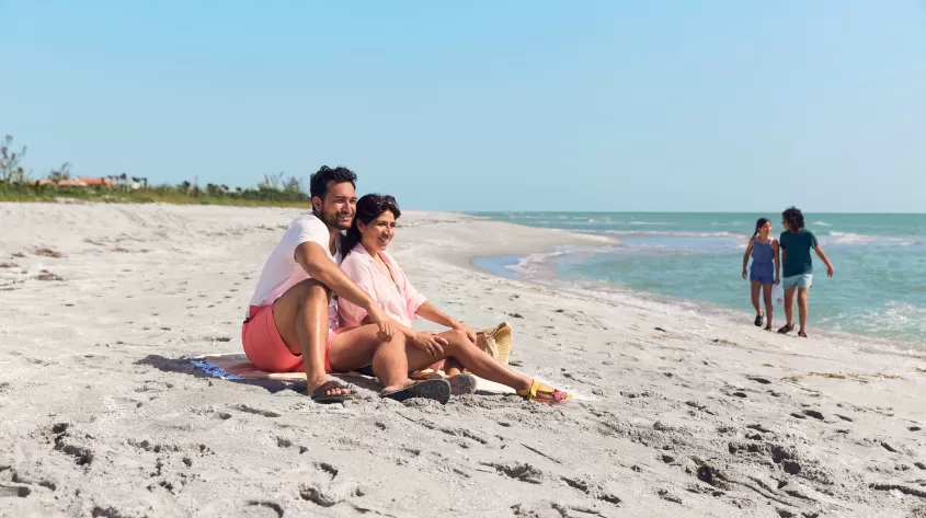 A couple sitting on top of a sandy beach with children in background