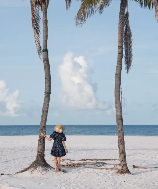 Back view of a woman in a dark floral dress and straw hat standing between two tall palm trees on a pristine white beach, looking out at the calm sea under a pale blue sky.